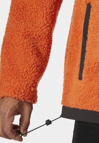 Oranžová fleecová bunda s texturovaným povrchem a černým akcentem na lemu. Nastavitelná šňůrka ve spodní části, viditelné ruce ji držící.
