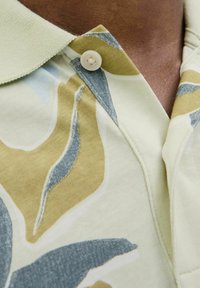 Camisa polo de manga corta en verde claro con patrones abstractos en beige, azul y gris. Presenta un cuello abotonado y una textura suave.