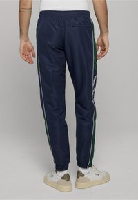 Granatowe spodnie dresowe z zielonymi bocznymi panelami, białym pipingiem i elastycznym pasem. Posiadają tylnią kieszeń i dopasowany, wąski krój.