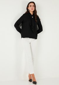 Black blouse with a bow tie detail at the collar, featuring long sleeves and button accents, paired with white fitted pants and black heels.