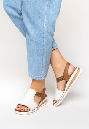 Füße in weißen und braunen Sandalen mit offenen Zehen und elastischen Riemen, kombiniert mit hellblauen Jeans, vor einem einfarbigen Hintergrund.