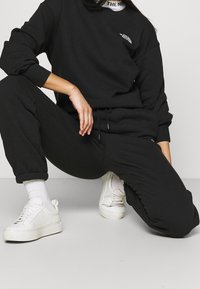 Schwarzer Baumwoll-Sweatshirt und Jogginghose, mit gerippten Bündchen, Rundhalsausschnitt und kleinem Logo. Kombiniert mit weißen Sneakers und Knöchelsocken.