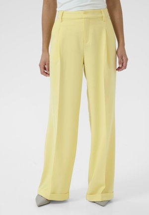 Osoba ubrana w żółte spodnie z wysokim stanem, szerokimi nogawkami i mankietami oraz szare szpiczaste buty, stojąca na jednolitym białym tle.