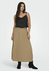 Top de satén negro con tirantes finos, combinado con una falda larga plisada beige. La modelo lleva botas negras de tobillo.