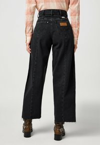 Jeans anchos negros con cintura alta, dobladillo deshilachado y bolsillos traseros. El material es denim con una textura sutil.