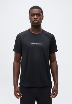 Jonge zwarte man die recht vooruit staat, draagt een zwart Peak Performance T-shirt en zwarte korte broek tegen een effen lichte achtergrond.