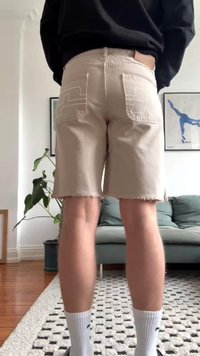 Bordinės smėlio spalvos džinsinės šortai su susidėvėjusiu kraštu, dvi galinės kišenės su siuvinėjimo dizainu, dėvimi su baltomis kojinėmis. Už jų matyti augalas ir sofa.