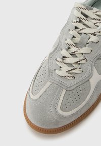 Szürke velúr sportcipő perforált részletekkel és krémszínű kiemelésekkel. Bézs színű fűzőkkel és texturált gumi talppal rendelkezik.