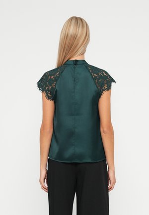 Kvinde med langt blond hår iført en mørkegrøn satin top med blonder korte ærmer, set bagfra, kombineret med sorte bukser.