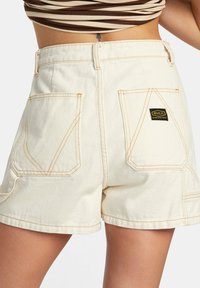 Beige jeanshorts med hög midja, med gula sömmar, två bakfickor och en svart logotypflik.