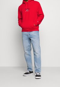 Felpa rossa con cappuccio e tasca frontale, con logo "Tommy Jeans New York City". Abbinata a jeans azzurri e sneakers nere.