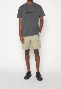 Grijze katoenen t-shirt met een reliëf 'carhartt.' logo, gecombineerd met beige cargoshorts voorzien van een zijzak, afgemaakt met zwarte instappers.