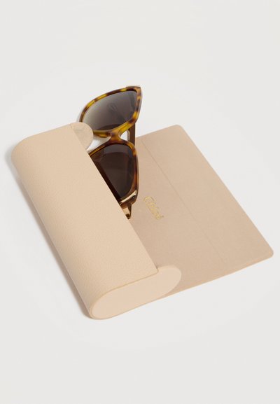 Γυαλιά ηλίου με καμήλια και μαύρα πλαίσια, εν μέρει μέσα σε μπεζ υφασμάτινη θήκη με την επιγραφή "Chloé" σε χρυσό στο ανοιχτό καπάκι.