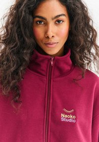 Bordowa bluza na zamek z wysokim kołnierzem, miękką fakturą oraz wyhaftowanym logo „Naoko Studio” z żółtym i wielokolorowym zakrzywionym akcentem.