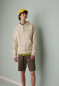 Sweatshirt beige à capuche avec poche avant et texte, superposé sur une chemise verte à motifs, associé à un short marron et une casquette jaune.