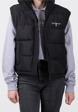 Person iført en sort dunvest over en lys lilla Calvin Klein hættetrøje og mørkegrå jeans, stående mod en ensfarvet baggrund.