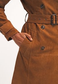 Brązowy zamszowy płaszcz trench z podwójnymi guzikami, paskiem w talii i bocznymi kieszeniami. Charakteryzuje się miękką teksturą i dopasowanym krojem.