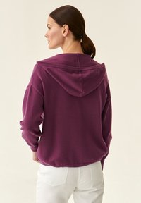 Felpa violacea con una vestibilità morbida, dotata di cappuccio con cordino e polsini a costine. Tessuto morbido, indossata sopra pantaloni bianchi.