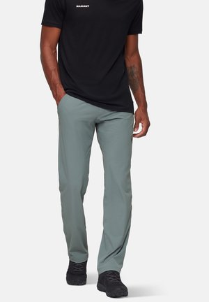 Helle graue Outdoor-Hosen mit lockerer Passform, aus atmungsaktivem Stoff. Kombiniert mit einem schwarzen T-Shirt und schwarzen Wanderschuhen.
