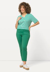 Maglietta a righe verdi e bianche con maniche corte e scollatura a V, abbinata a pantaloni verdi aderenti e scarpe bianche con lacci su uno sfondo neutro.