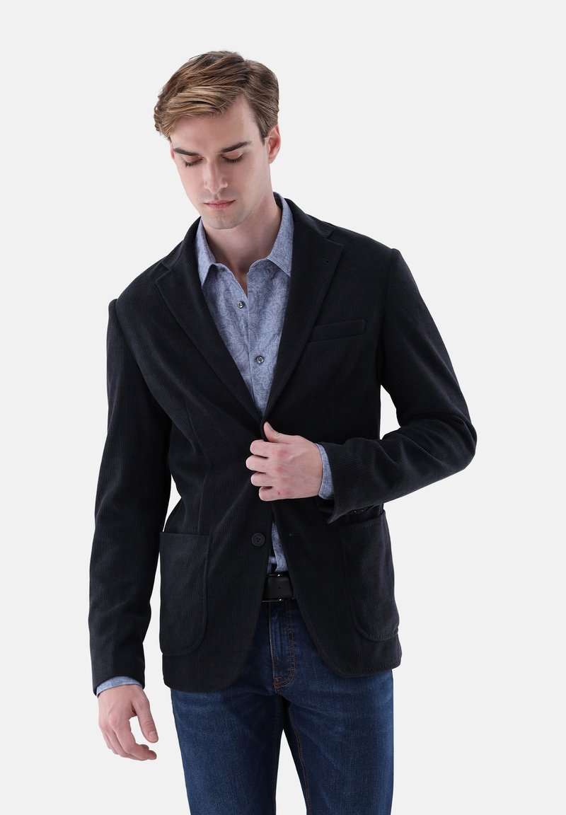 Schwarzer Cord-Blazer mit Reverskragen, einer Brusttasche und zwei Vordertaschen, getragen über einem hellblauen, gemusterten Hemd und dunklen Jeans.