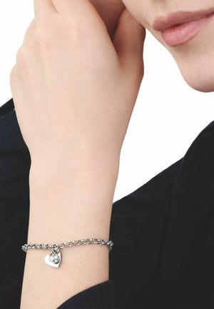 Zilveren armband met een hartvormige bedel en een heldere edelsteen, gedragen om een pols tegen een zwarte achtergrond met een lange mouw.