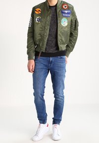 Veste bomber vert olive avec des patchs, fermeture éclair, poignets côtelés, portée par-dessus un pull sombre. Associée à un jean bleu et des baskets blanches.