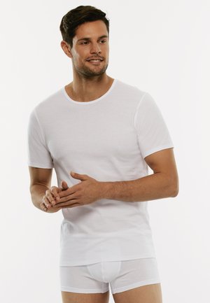 T-shirt bianco a maniche corte in morbido tessuto, con scollo rotondo. Indossato con boxer aderenti bianchi. Il modello mostra una posa rilassata con le mani unite.