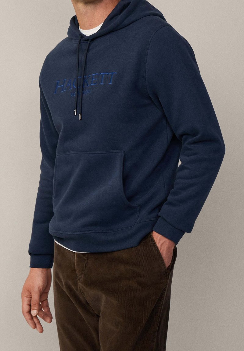 Homme portant un sweat à capuche Hackett bleu marine avec poche avant et pantalon en velours côtelé marron, main dans la poche, fond neutre.