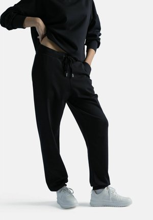 Henkilö, joka pukeutunut mustiin nyöripanta housuihin ja mustaan pitkähihainen toppiin, valkoiset lenkkarit jalassa, toinen käsi taskussa ja toinen lanteilla.