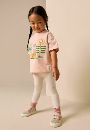 Fiatal lány fonott hajjal, rózsaszín virágmintás pólót, világos leggingset, rózsaszín zoknit és pasztell színű tornacipőt viselve, beltéren áll.