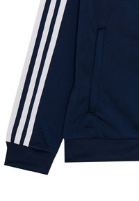 Námořnicky modrá bunda s třemi bílými pruhy podél rukávu. Má měkký, strukturovaný povrch a kapsu na boku u lemu.