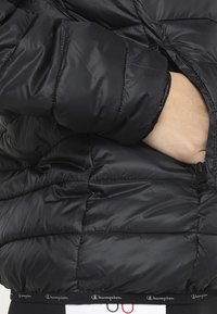 Svart pufferjacka med kviltat mönster, dragkedjeficka och elastiska muddar. Har en logotypdetalj på mudden och nederkanten.