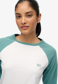 Witte en teal lange mouw raglan shirt met een ronde hals. Zacht katoenmateriaal. Heeft het "Super Dry" logo in lichtblauw op de borst.