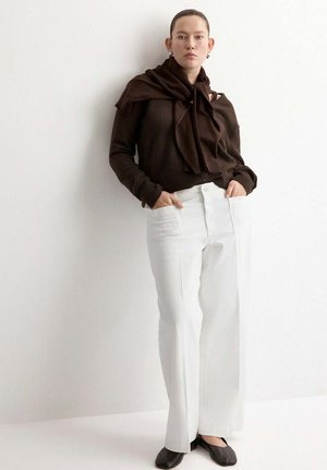 Mujer de pie con las manos en los bolsillos de un pantalón blanco de cintura alta, vistiendo una blusa de manga larga marrón oscuro y un suéter a juego sobre los hombros.