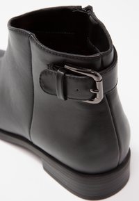 Gros plan sur une botte noire en cuir avec une lanière à boucle argentée autour de l'arrière et des détails cousus sur une surface lisse.