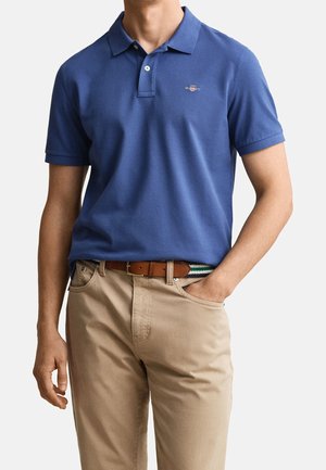 Uomo con polo blu, pantaloni beige e cintura marrone, in piedi con una mano in tasca su uno sfondo chiaro e uniforme.