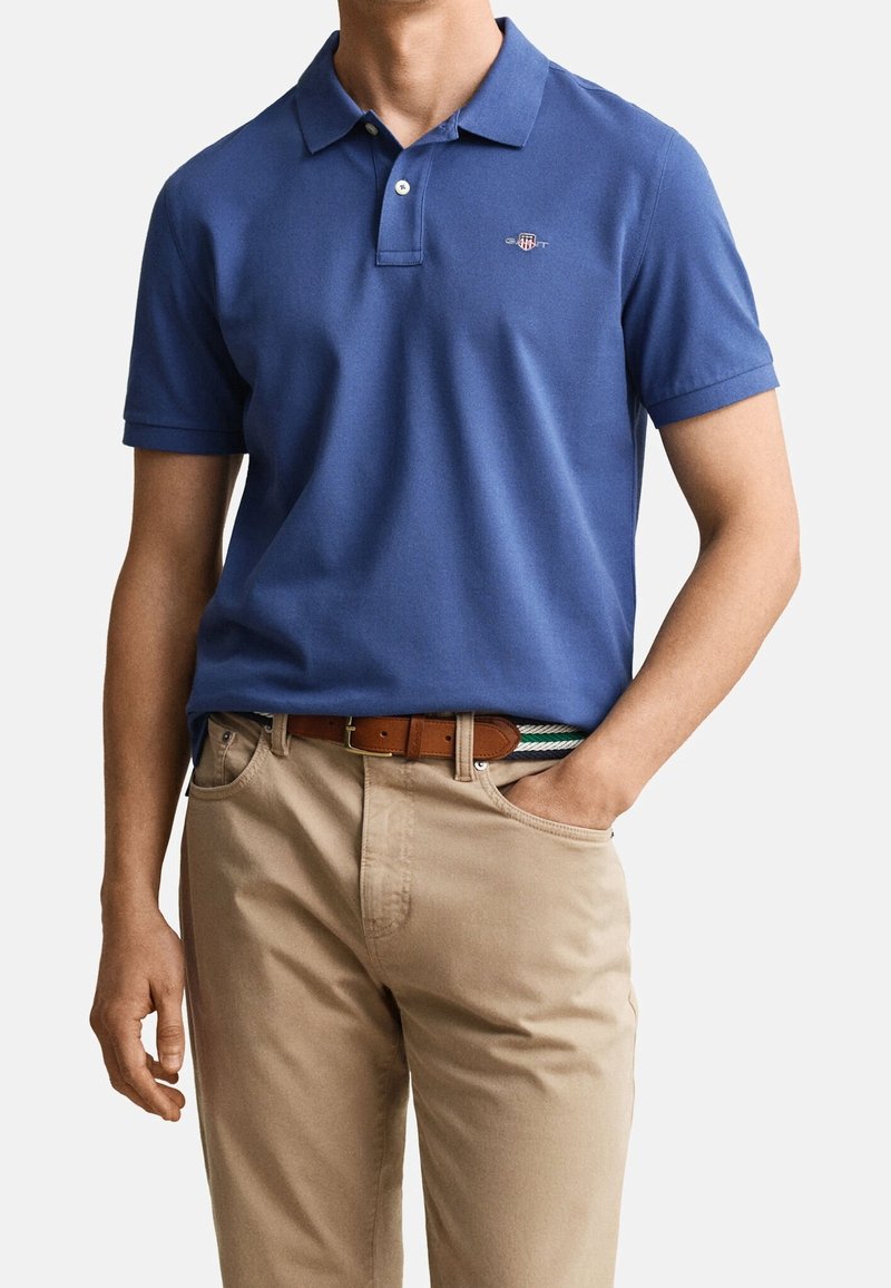 Homme portant un polo bleu, un pantalon beige et une ceinture marron, debout avec une main dans la poche sur un fond uni clair.