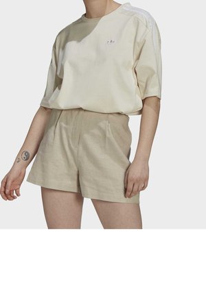 Osoba nosząca beżowe szorty z wysokim stanem oraz luźną kremową koszulkę Adidas z logo i paskami na ramionach, pokazująca tatuaż yin-yang na lewym przedramieniu.
