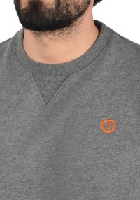 Pelēka džemperis ar ribotu apaļu kakla izgriezumu un oranžu apļa formālu logotipu. Audums izskatās mīksts ar viegli teksturētu virsmu.