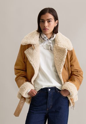 Bruine schapenvachtjas met een pluizige crèmekleurige kraag en manchetten, gedragen over een wit shirt en donkere spijkerbroeken. Zilveren hardwareaccenten.