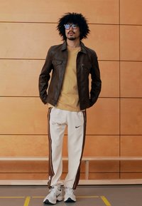 Καφέ δερμάτινο μπουφάν και μουσταρδί μπλουζάκι, λευκή φόρμα με καφέ ρίγες και λογότυπο Nike, αθλητικά παπούτσια, με σγουρά μαύρα μαλλιά και γυαλιά.