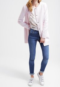 Ljusrosa lång cardigan med rund hals och knappstängning, bärs över en mönstrad topp, tillsammans med åtsittande blå jeans och vita sneakers.