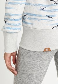 Sudadera gris con rayas azules y estampados de pájaros. Presenta puños y dobladillo acanalados, con una etiqueta de logo en el borde inferior. Leggings de tela gris visibles.
