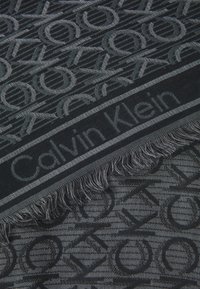 Mustavalkoinen kuvioitu huivi, jossa on kudottu Calvin Klein -logo, frangeilla varustettu reuna ja sileä pinta.