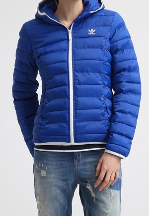 Veste matelassée Adidas bleue à capuche avec fermeture éclair blanche et logo portée par une personne en jean bleu usé, main reposant près de la poche.