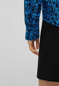 Kvinnas hand bredvid en svart kjol, bär en långärmad tröja med blå och svarta abstrakta mönster och knäppta manschetter.