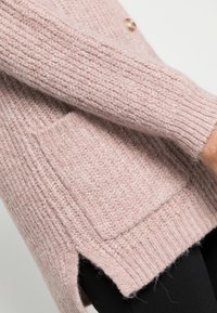 Dzianinowy sweter zapinany na guziki w delikatnym różu z teksturowanym wzorem, z przednią kieszenią, ściągaczami przy rękawach i bocznymi rozcięciami dla większej swobody ruchów.