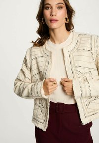 Giacca beige con texture, adornata da embellishments geometrici, bordi frangiati e rifiniture metalliche, indossata sopra un top crema e pantaloni bordeaux.