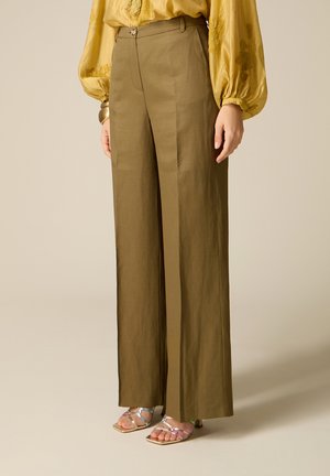Frau trägt olivgrüne Hosen mit weitem Bein, gelbe langärmlige Bluse mit Stickerei, metallische Sandalen mit offenen Zehen und goldenes Armband.